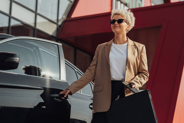 Elegante vrouwelijke ondernemer met koffer in de hand Opening deur van de luxe auto - Foto, afbeelding