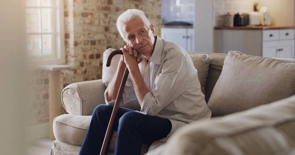Where did all the time go. Shot of a senior man sitting alone on the sofa at home and looking contemplative while holding his walking stick. - Photo, image