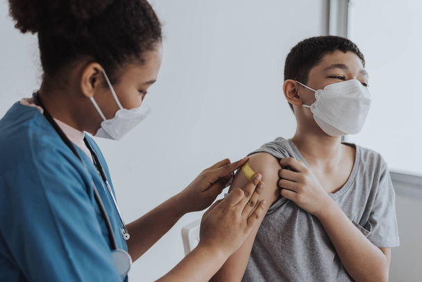 Ασιάτης με μάσκα προσώπου που δέχεται εμβόλια από γιατρό. Ένα παιδί εμβολιάζεται από γιατρό και νοσοκόμα για να αποτρέψει τον ιό της στέψης. Εκστρατεία εμβολιασμού Covid-19 σε κλινική. - Φωτογραφία, εικόνα