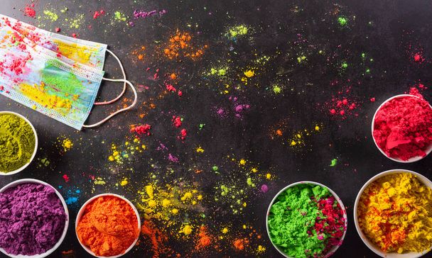 Vista superior de polvo de holi colorido con pintura holi máscara médica multicolor sobre fondo oscuro. Feliz festival de Holi durante coronavirus - Foto, imagen