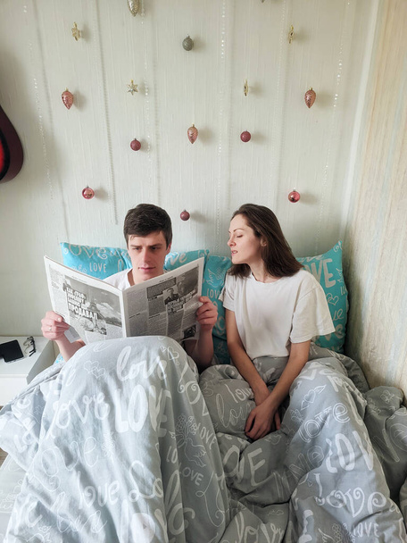Happy couple lying in bed in cozy home, drinking coffee. Young man in a t-shirt and a cute woman. Family holiday and togetherness, love. Weekend. Lifestyle. - 写真・画像