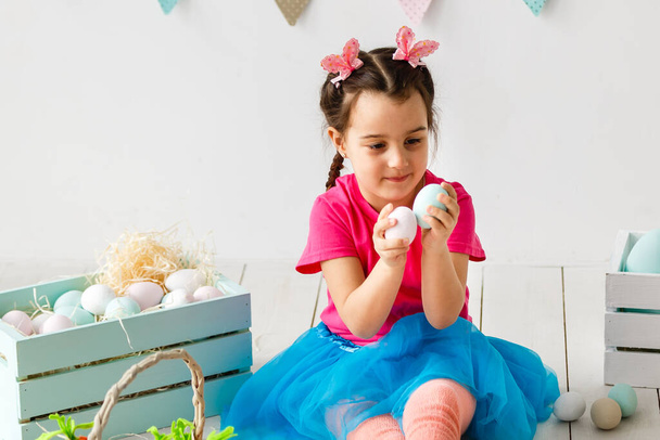 Готовлюсь к Пасхе. Милая маленькая девочка держит пасхальное яйцо и улыбается с украшением на заднем плане - Фото, изображение