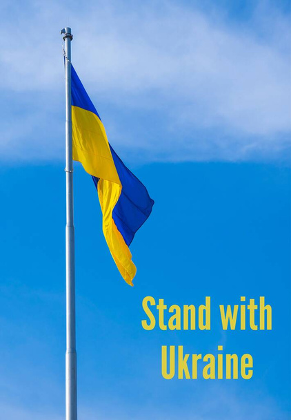 STAND WITH UKRAINE text on Ukrainian national flag flutters in the wind against the blue sky. National symbol of ukrainian people - blue and yellow. Independence Praying, mourning, humanity. No war. War Protest against Russian invasion of Ukraine. - Photo, Image