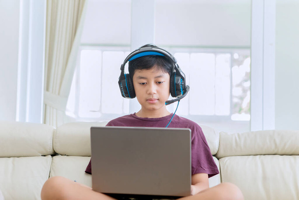 Preteen boy looks bored while using a laptop and headphones on the sofa. Shot at home - Valokuva, kuva