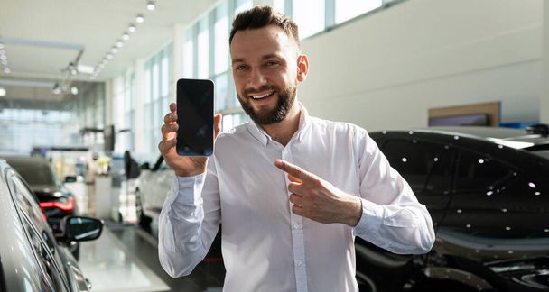 egy férfi egy autókereskedésben mosollyal mutatja be az okostelefon képernyőjét - Fotó, kép