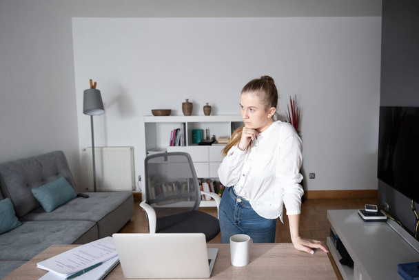 Werken vanuit huis en freelancen concept. Jonge vrouw staan en denken op project in de voorkant van laptop. - Foto, afbeelding