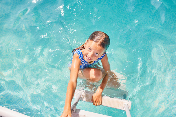Ein Kind auf der Treppe, das aus dem Pool klettert, kleines Mädchen, das Spaß im Schwimmbad hat, Sommerurlaub zu Hause, tropischer Ferienort. - Foto, Bild