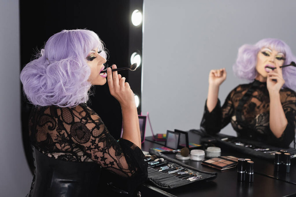 Aynanın yanında kozmetik fırçasıyla oturan eksantrik travesti - Fotoğraf, Görsel