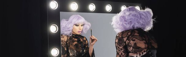 трансвестит в фиолетовом парике, держа косметическую кисть, глядя в зеркало, баннер - Фото, изображение