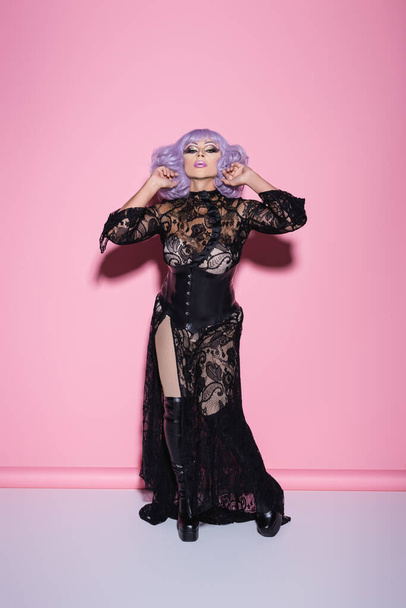 full length view of eccentric drag queen in black lace dress looking at camera while standing on pink - Photo, Image