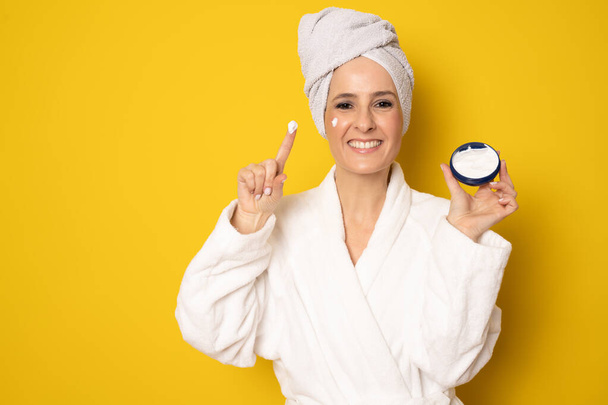 Happy young woman in bathrobe with cream in her face standing isolated over yellow background. Beauty concept. - Foto, Bild