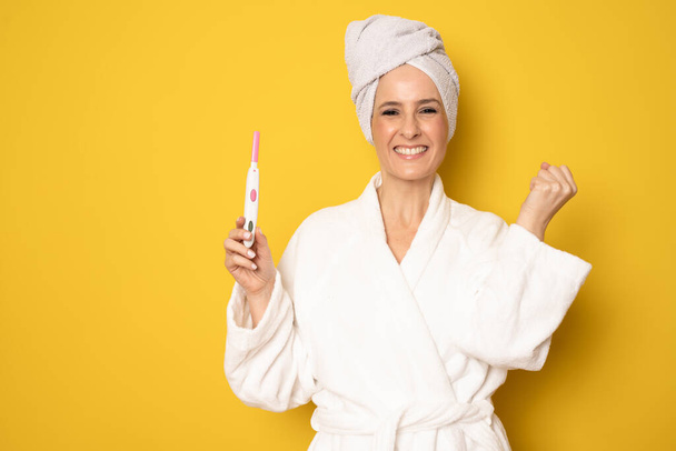 Happy woman in bathrobe holding pregnancy test standing isolated over yellow background. - Fotografie, Obrázek