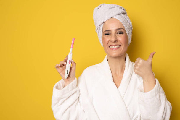 Happy woman in bathrobe holding pregnancy test standing isolated over yellow background. - Foto, Imagen