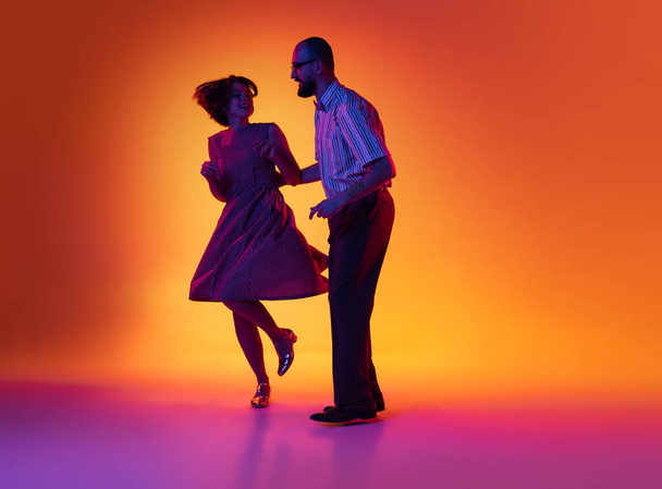 Portrait of excited man and woman, couple of dancers in vintage retro style outfits dancing lindy hop dance isolated on gradient yellow and purple background. - Fotó, kép