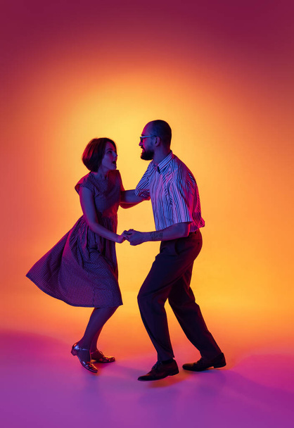 Portrait of excited man and woman, couple of dancers in vintage retro style outfits dancing lindy hop dance isolated on gradient yellow and purple background. - Φωτογραφία, εικόνα