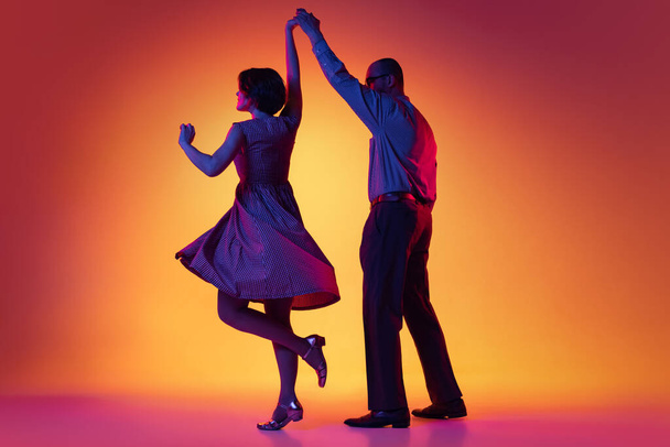 Portrait of excited man and woman, couple of dancers in vintage retro style outfits dancing lindy hop dance isolated on gradient yellow and purple background. - Foto, afbeelding