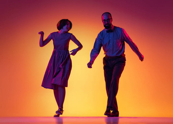 Portrait of excited man and woman, couple of dancers in vintage retro style outfits dancing lindy hop dance isolated on gradient yellow and purple background. - Foto, imagen