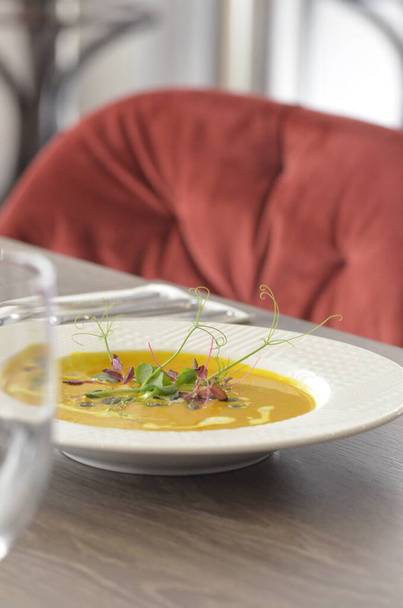 суп из желтых сливок с тыквой масла с семенами и сливками, дополненный свежими цветными микротравами, на белой тарелке и деревянном столе, фотографии обеденного меню - Фото, изображение