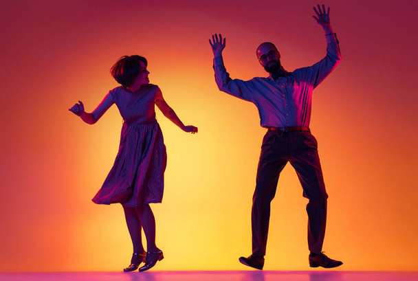 Portrait of excited man and woman, couple of dancers in vintage retro style outfits dancing lindy hop dance isolated on gradient yellow and purple background. - Фото, изображение