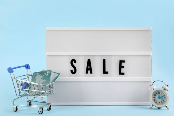 Lightbox with word Sale, alarm clock and credit cards in shopping cart on light blue background - Fotó, kép