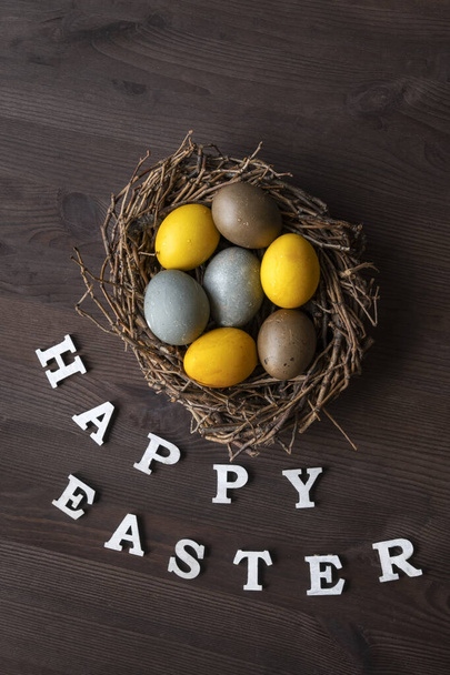 Vista superior de la inscripción Feliz Pascua y huevos de colores en un nido de aves en una mesa de madera marrón. Marco vertical. - Foto, Imagen