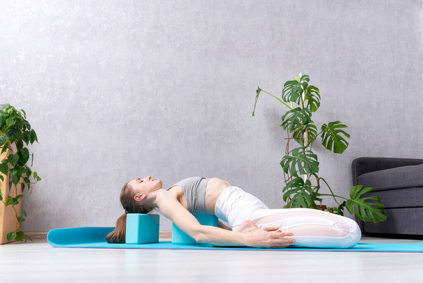 Young woman in white sportwear practicing yoga indoors at home. Exercises with yoga blocks. Home workout. - Foto, Imagen