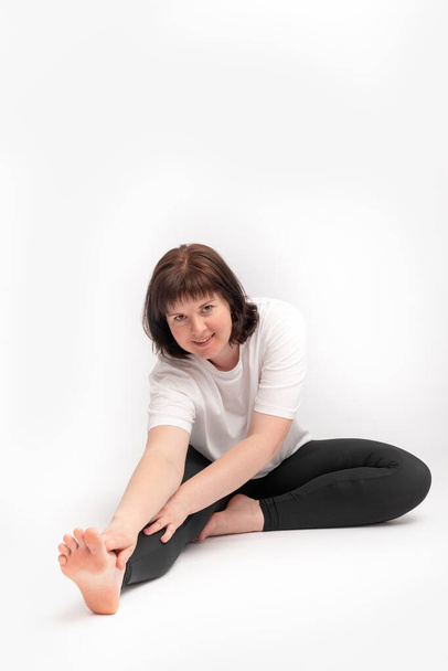 Plus size young brunette woman doing stretching exercise and looks at the camera. Portrait of girl on white background engaged in gymnastics - Photo, Image