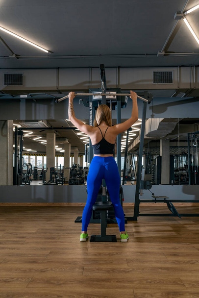 Jeune femme mince en legging serré s'entraîne dans la salle de gym. Vue de derrière. Poussée du bloc supérieur. Cadre vertical - Photo, image