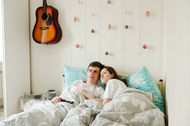 Egy boldog pár fekszik az ágyban egy otthonban, kávézgatnak. Fiatal férfi pólóban és egy csinos nővel. Családi nyaralás és együttlét, szerelem. Hétvége. Életmód. - Fotó, kép