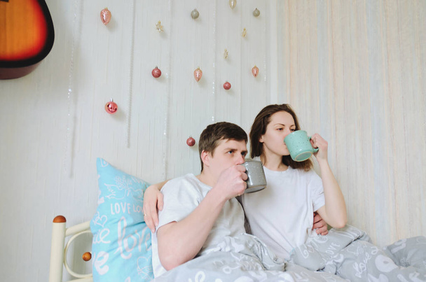 Ευτυχισμένο ζευγάρι ξαπλωμένο στο κρεβάτι στο άνετο σπίτι, πίνοντας καφέ. Νεαρός με μπλουζάκι και χαριτωμένη γυναίκα. Οικογενειακές διακοπές και συντροφικότητα, αγάπη. Σ / Κ. Τρόπος ζωής. - Φωτογραφία, εικόνα