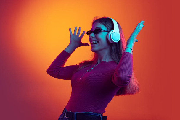 Portrait of young beautiful happy girl in headphones listening to music isolated on orange background in neon light, filter. Concept of emotions, music - Photo, Image