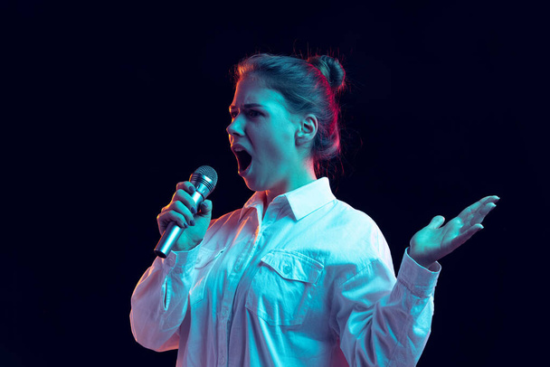 Half-length portrait of young pretty girl in white shirt with microphone isolated on dark background in blue neon light, filter. Concept of emotions, fashion, youth - Foto, Imagen