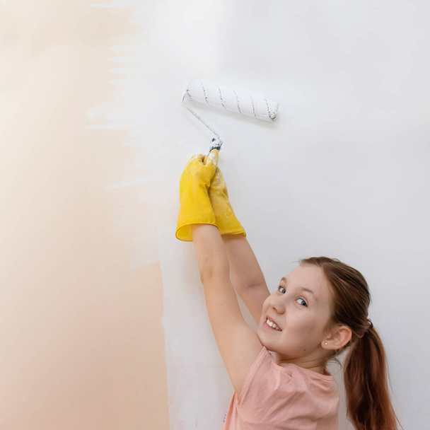 renovación de la casa. pintura de paredes con paint.a chica en guantes amarillos pinta una pared con un rodillo de pintura blanca y smiles.selective foco - Foto, imagen