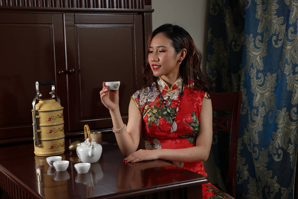 Aantrekkelijke jonge vrouw dragen retro Chinese cheongsam jurk zitten naast vintage tafel met thee set tiffin carrier stoel kijken holding controleren poseren pour drankje aanbod thee - Foto, afbeelding