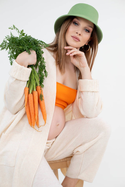 Verticale schoonheid zwangere vrouw groene hoed, oranje top en vlas kleding, naakt buik op zoek camera, sinaasappel wortelen. - Foto, afbeelding