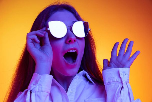 Portrait of young beautiful emotive girl in white shirt isolated on orange background in neon light, filter. Concept of emotions, fashion, youth - Φωτογραφία, εικόνα
