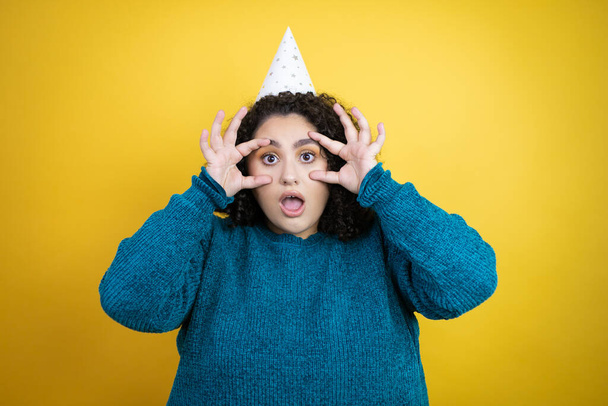 Young beautiful woman wearing a birthday hat over isolated yellow background Trying to open eyes with fingers, sleepy and tired for morning fatigue - Photo, Image