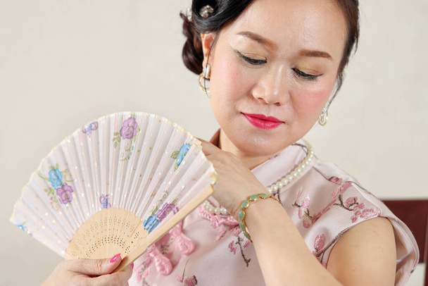 Aantrekkelijke vrouw van middelbare leeftijd het dragen van Chinese cheongsam jurk zitten op stoel met poseren Chinese versierde hand ventilator - Foto, afbeelding