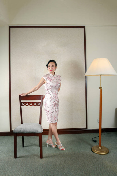 Приваблива жінка середнього віку, одягнена в китайську сукню Чонсам, стоїть поруч з кріслом і підлоговою лампою. - Фото, зображення