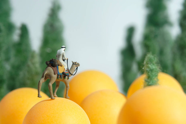 Kreatywne miniaturowe ludzie zabawka fotografia figura. Mężczyźni w białych ciuchach kandury jadący na wielbłądzie nad żółtą kulą na jasnym niebie. Zdjęcie obrazkowe - Zdjęcie, obraz