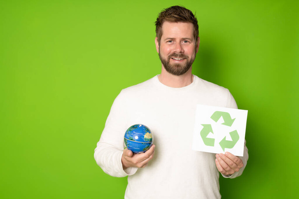 Joven hombre caucásico guapo aislado sobre fondo verde sosteniendo un cartel con icono de reciclaje y tierra globo. - Foto, imagen