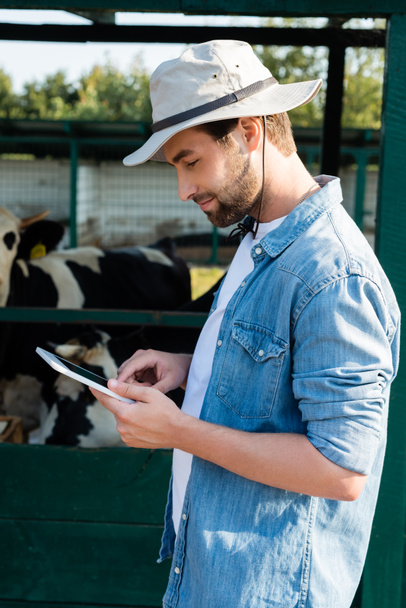 vista lateral del granjero sonriente en sombrero de ala utilizando tableta digital cerca de establo en la granja - Foto, Imagen