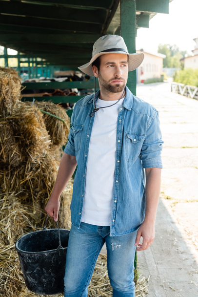 Jungbauer in Jeanshemd und Krempenhut hält Eimer in der Hand und schaut auf Bauernhof weg - Foto, Bild
