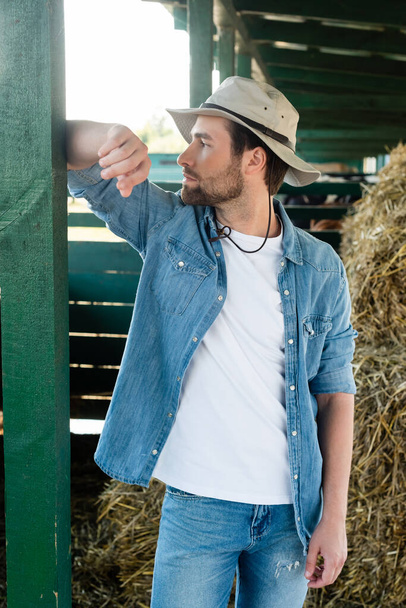 farm worker in denim shirt and brim hat looking away while standing near barn - Фото, изображение