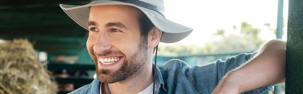 portrét mladého vousatého farmáře v klobouku usmívajícího se na statku, prapor - Fotografie, Obrázek
