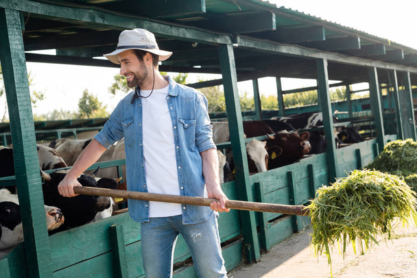 alegre granjero en ala sombrero sosteniendo heno cerca de la casa de vaca en la granja - Foto, Imagen