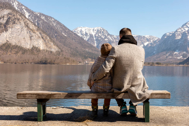 Ojciec i syn siedzą razem na ławce i cieszą się górami, śniegiem, dobrą pogodą, błękitnym niebem. Mały chłopiec i jego ojciec spędzają razem czas. Rodzinny czas. Piękny zimowy krajobraz. - Zdjęcie, obraz