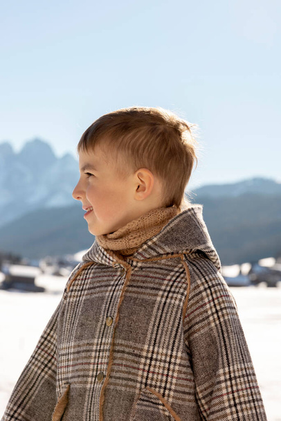 Portrait of little adorable boy with winter landscape in the background. Cute, smiling caucasian child with brown coat standing outdoor and enjoying good weather, blue sky, snow. Good, positive mood. - Фото, изображение