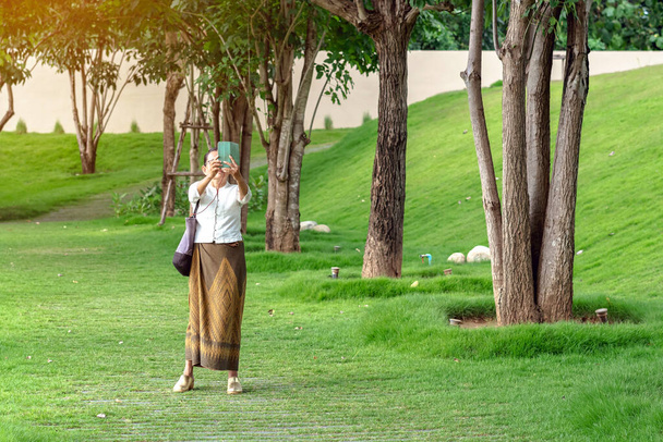 Ázsiai női séta és használja okostelefon, hogy egy szelfit a park ösvényén magas zöld fák. Egy nő mobilt használ, hogy képeket készítsen, miközben a parkban pihen. Boldog emberek a zöld természetben. - Fotó, kép