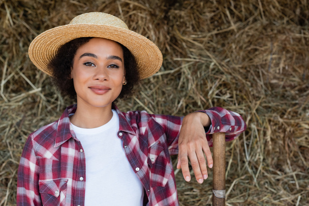 positive african american woman in plaid shirt and straw hat smiling at camera near blurred haystack - Φωτογραφία, εικόνα